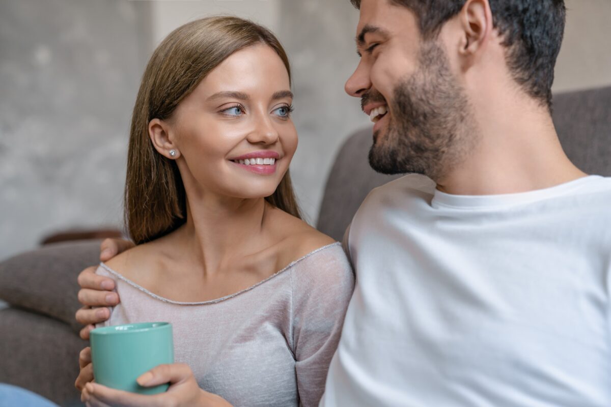 Young cheerful couple sitting together on couch in living room drinking coffee and talking