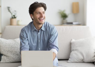 Video Call Concept. Cheerful Man In Wireless Earphones Using Laptop Communicating With Friends Sitting On Couch At Home.
